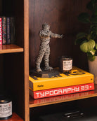 String Thing® guitar string upcycling mummy displayed as music room shelf art — fully wrapped with used guitar strings alongside FRET12 candle and design books and cassette tapes